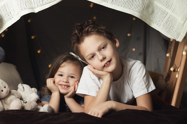 Geschwister liegen in einer Hütte mit Stühlen und Decken. Bruder und Schwester lächeln und spielen zu Hause