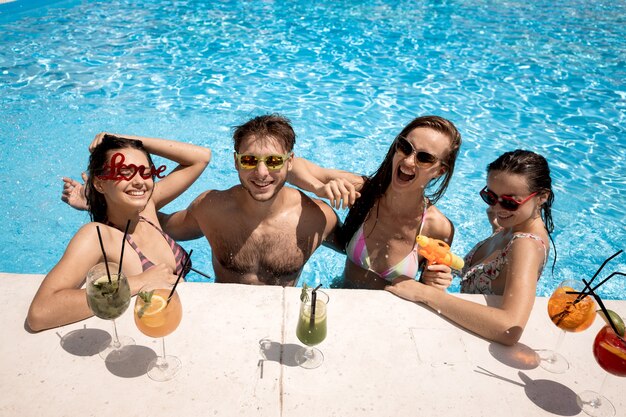 Gesellschaft junger fröhlicher Mädchen und Kerle, die sich an einem sonnigen Sommertag neben der Lounge-Zone im Pool entspannen, Cocktails trinken und mit Wasserpistolen im Freien spielen