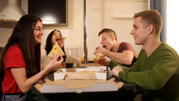 Gesellschaft von vier jungen Freunden, die in der Küche sitzen und Pizza essen
