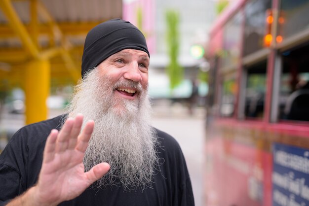 Gesicht des glücklichen reifen gutaussehenden bärtigen Hipster-Mannes, der Hand am Bus winkt