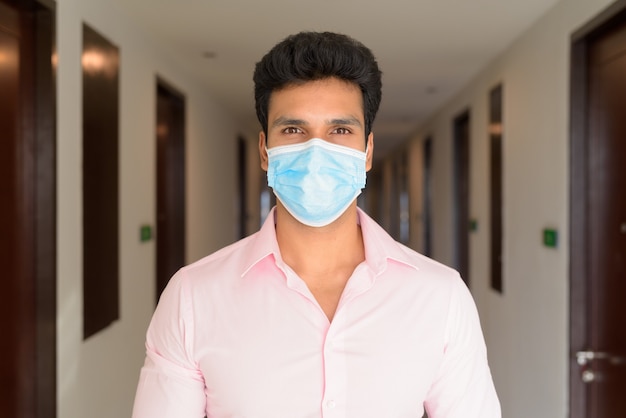 Gesicht des jungen indischen Geschäftsmannes, der Maske zum Schutz vor Ausbruch des Koronavirus im Korridor trägt