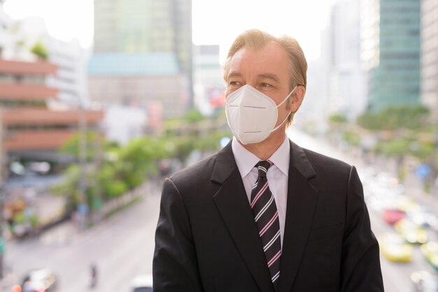 Gesicht des reifen Geschäftsmannes mit Maske, die auf Ansicht der Stadt denkt