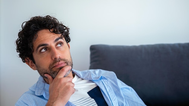 Gesicht eines nachdenklichen erwachsenen Mannes, der nachdenklich aufschaut, während er auf der Couch zu Hause sitzt. Bärtiger Mann, der die Wahl trifft oder auf einem Sofa denkt, das wegschaut. Eine Person auf weißem Hintergrund