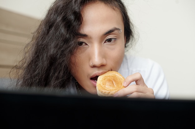 Gesicht eines nachdenklichen jungen Mannes mit langen lockigen Haaren, der süßes hausgemachtes Gebäck isst und einen interessanten Artikel auf dem Laptop-Bildschirm liest