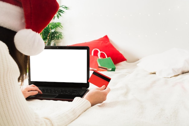 Gesichtslose Frau in der Weihnachtsmannmütze, die zu Hause mit Laptop und Plastikkarte auf Bett liegt
