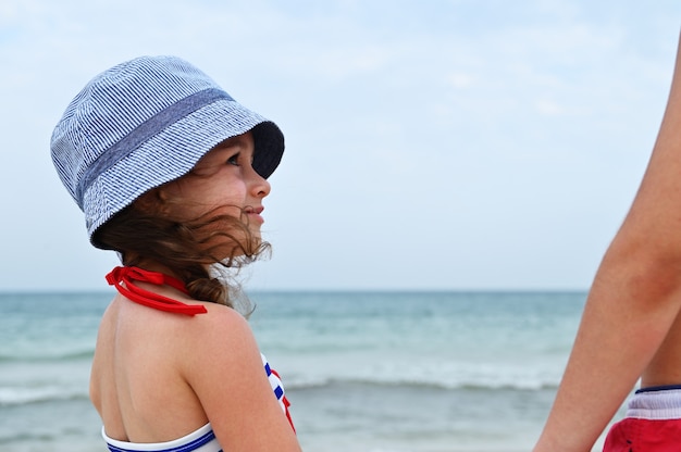 Gesichtsporträt des schönen kleinen Mädchens in Sommerhut und Badeanzug, das zur Seite schaut, Sommerferien auf See genießt und gesundes Sonnenbaden im Freien nimmt. Landschaftsseelandschaft.