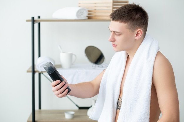 Gesichtsreinigung Hautbehandlung Morgenroutine Schöner fröhlicher junger Mann weißes Handtuch auf den Schultern mit Schaumflasche im hellen Duschraum