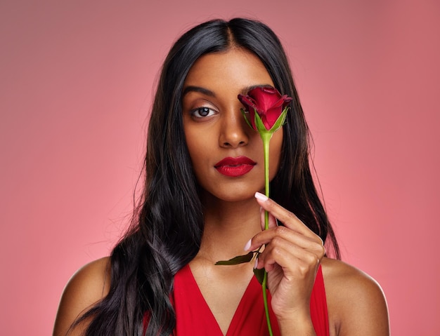 Gesichtsschönheit und Porträt einer Frau mit einer Rose auf einem Studiohintergrund für den Valentinstag. Make-up-Modell und ernstes junges indisches Mädchen mit einer Blume in der Hand für Romantik oder Liebe auf rosa Hintergrund