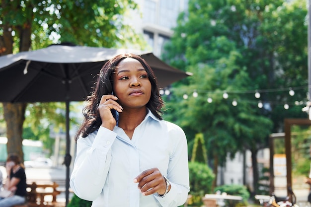 Gespräch per Telefon führen Junge afroamerikanische Frau in weißem Hemd im Freien in der Stadt in der Nähe von grünen Bäumen und gegen Geschäftsgebäude