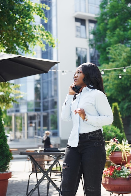 Gespräch per Telefon führen Junge afroamerikanische Frau in weißem Hemd im Freien in der Stadt in der Nähe von grünen Bäumen und gegen Geschäftsgebäude