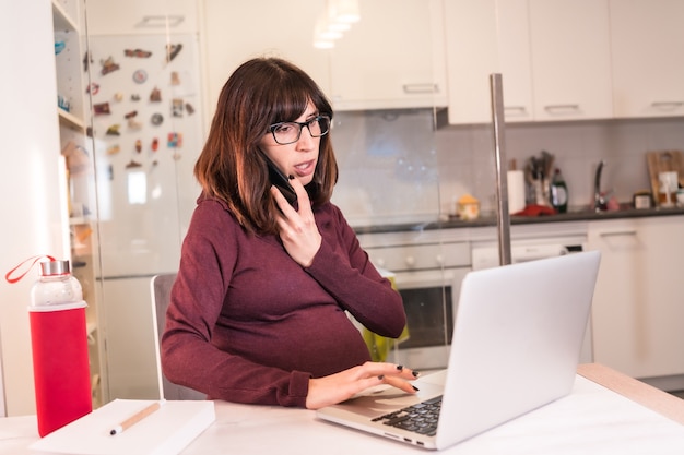 Gestante jovem fazendo teletrabalho de casa no computador devido às dificuldades de trabalho, fazendo ligação para o trabalho