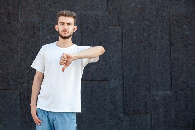 Gesten-Emotionsausdruck und Personenkonzept Europäischer bärtiger, finsterer Mann in weißem T-Shirt und Jeans, der mit der linken Hand draußen auf dunklem Hintergrund mit Kopierraum Daumen nach unten zeigt