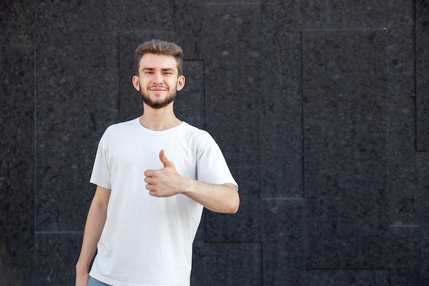 Gesten-Emotionsausdruck und Personenkonzept Europäischer bärtiger glücklicher dunkelhaariger Mann in weißem T-Shirt, das Daumen nach oben mit seiner linken Hand im Freien auf einem dunklen Hintergrund mit Kopierraum zeigt