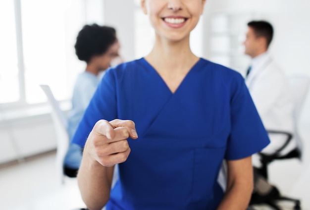 Gesten-, Menschen- und Medizinkonzept - Nahaufnahme einer glücklichen Ärztin oder Krankenschwester, die mit dem Finger auf Sie zeigt, während sich eine Gruppe von Medizinern im Krankenhaus trifft