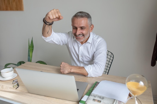 gesto amistoso. Sonriente hombre de mediana edad de cabello gris con el puño levantado mirando la pantalla del portátil sentado en la mesa en la oficina