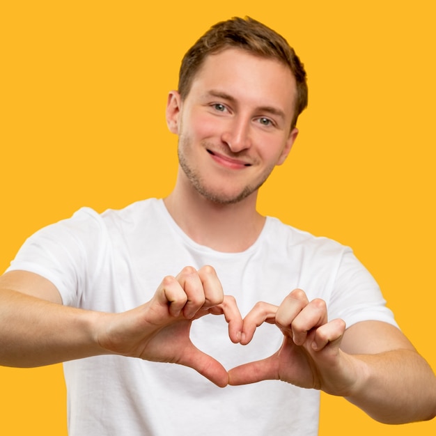 Gesto de corazón. Devoción al amor. Hombre feliz en camiseta blanca que muestra signo romántico aislado en la pared amarilla.