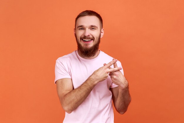 Foto gesto de hashtag retrato de un hombre barbudo cruzando los dedos mostrando el símbolo de hash etiquetando un famoso mensaje de internet blog viral usando una camiseta rosa disparo de estudio interior aislado en fondo naranja