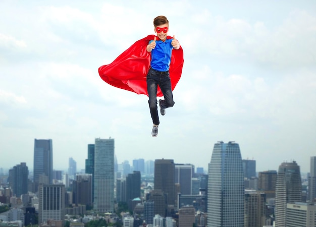 gesto, libertad, infancia, movimiento y concepto de personas - niño con capa de superhéroe rojo y máscara volando en el aire y mostrando los pulgares hacia arriba sobre el fondo de la ciudad