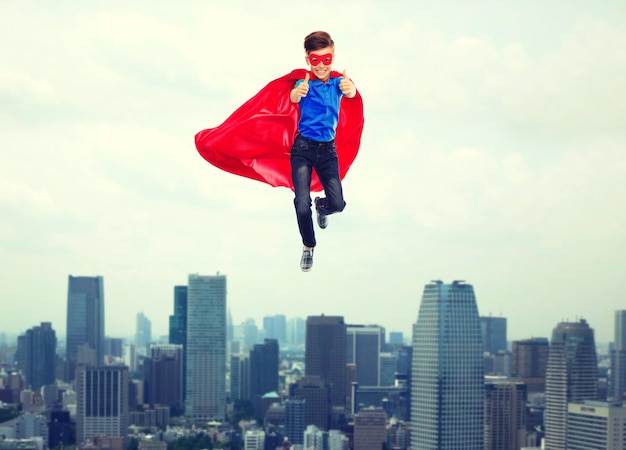 gesto, libertad, infancia, movimiento y concepto de personas - niño con capa de superhéroe rojo y máscara volando en el aire y mostrando los pulgares hacia arriba sobre el fondo de la ciudad