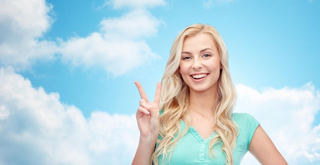 Gesto positivo y concepto de la gente: mujer joven sonriente o adolescente que muestra el signo de la mano de paz sobre el cielo azul y el fondo de las nubes