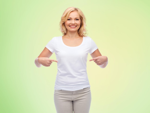 gesto, publicidad y concepto de la gente - mujer sonriente de mediana edad con una camiseta blanca en blanco apuntándose con el dedo a sí misma sobre un fondo natural verde