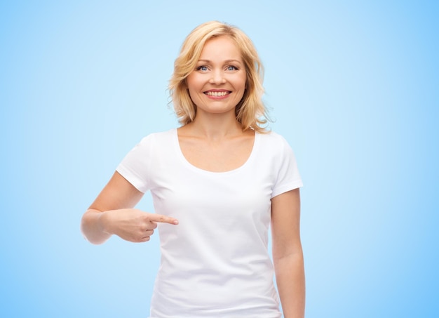 gesto, publicidad y concepto de la gente - mujer sonriente de mediana edad con una camiseta blanca en blanco apuntándose con el dedo sobre el fondo azul