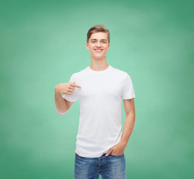 gesto, publicidad, educación, escuela y concepto de la gente - joven sonriente con una camiseta blanca en blanco apuntándose con el dedo sobre el fondo de la pizarra verde