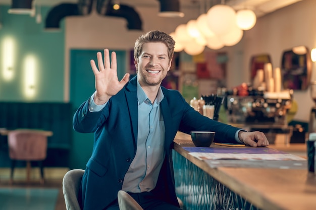 Gesto, saludo. Hombre atractivo sonriente en un traje de negocios que levantó la mano en señal de saludo mientras estaba sentado con un café en la cafetería