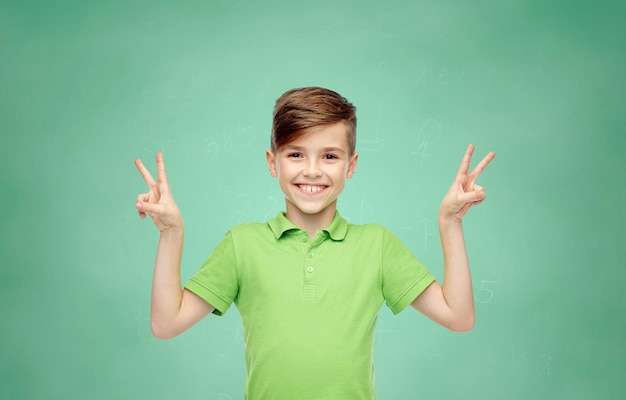 gesto, triunfo, infância, moda e conceito de pessoas - menino sorridente feliz em camiseta polo verde mostrando paz ou sinal de mão de vitória sobre o fundo do quadro de giz da escola verde