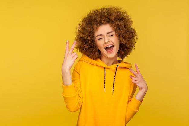 Gesto de victoria Retrato de una mujer entusiasta de pelo rizado con capucha de estilo urbano que muestra el signo v o la paz confiando en el éxito estudio de estilo de vida optimista aislado en fondo amarillo