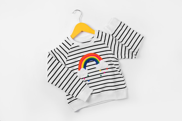 Gestreifter Pullover mit Regenbogen auf Aufhänger auf weißem Hintergrund. Süßes Kinderoutfit. Kinderkleidung für Herbst oder Frühling.