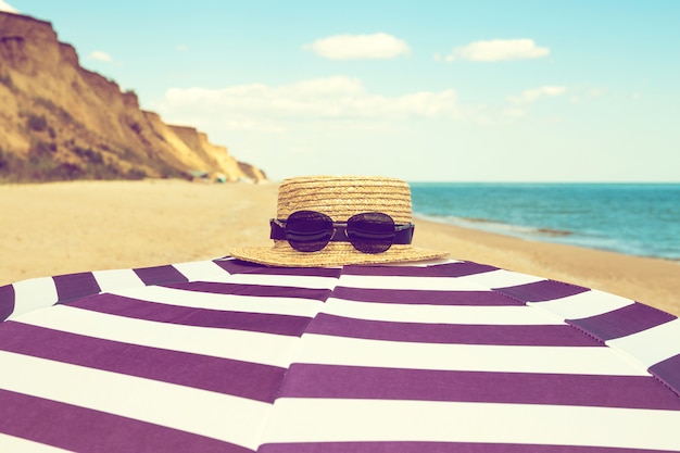 Gestreifter Strandschirm mit Strohhut und Sonnenbrille auf dem Seeufer mit Sand