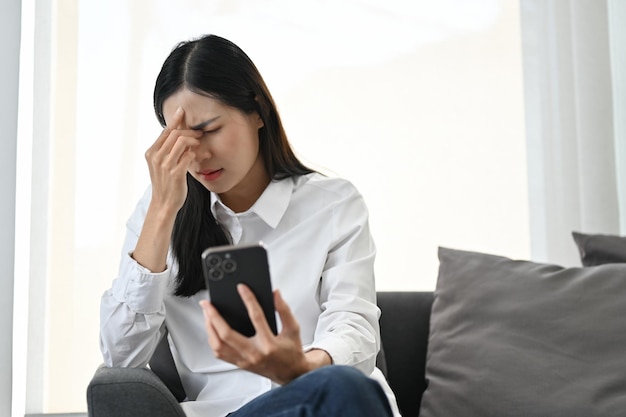 Gestresste asiatische Frau, die ihr Smartphone benutzt, während sie auf dem Sofa sitzt und unter Kopfschmerzen leidet