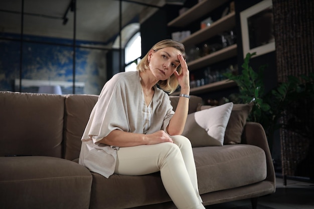 Gestresste Frau mittleren Alters sitzt gedankenverloren auf dem Sofa im Wohnzimmer.