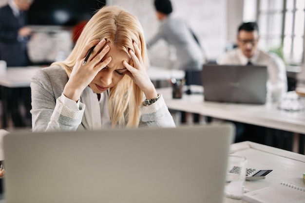 Gestresste Geschäftsfrau hat starke Kopfschmerzen, während sie an ihrem Schreibtisch im Büro arbeitet. Es gibt Menschen im Hintergrund