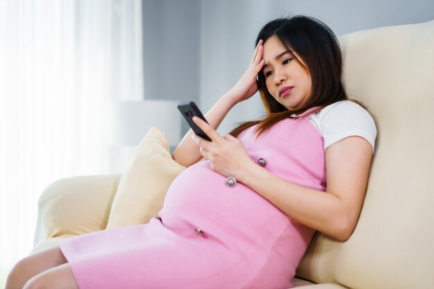 Gestresste schwangere Frau mit einem Smartphone auf Sofa im Wohnzimmer