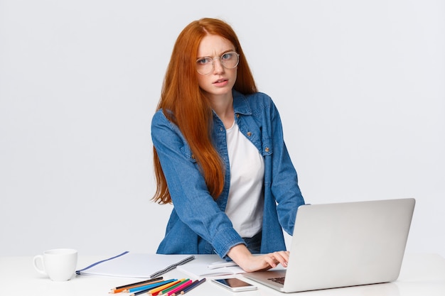 Gestresste und genervte, ernsthaft aussehende, rothaarige, fleißige Frau mit roten Haaren, stirnrunzelnd, sauer, mit viel Arbeit, mit Laptop und weißer Wand