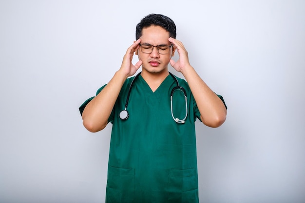 Gestresster asiatischer junger Arzt oder Krankenpfleger in grüner Uniform mit Stethoskop mit Kopfschmerzen