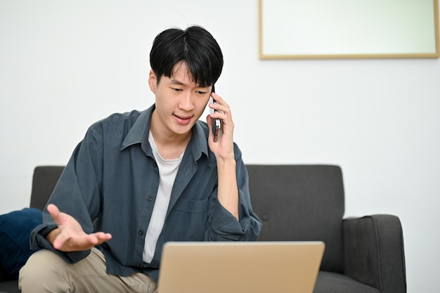 Gestresster asiatischer Mann, der auf den Laptop-Bildschirm schaut, während er ein ernstes Gespräch am Telefon führt