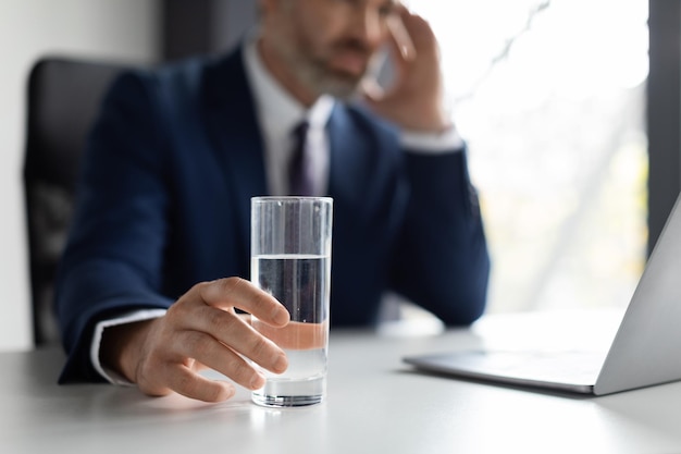 Gestresster Geschäftsmann, der am Arbeitsplatz unter Kopfschmerzen leidet und ein Glas Wasser hält