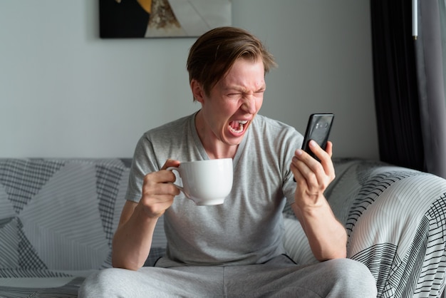 Gestresster junger Mann, der Kaffee hält, während er zu Hause am Telefon schreit