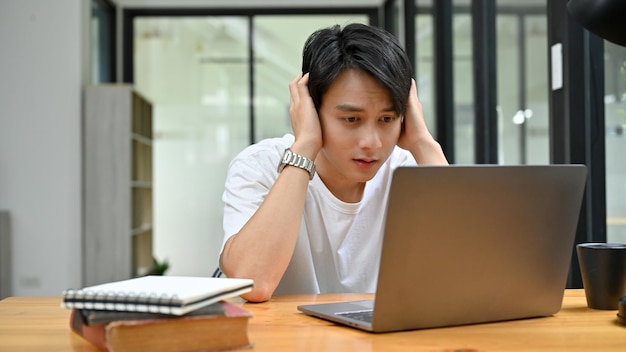 Gestresster und depressiver asiatischer männlicher Büroangestellter oder Student, der auf den Laptop-Bildschirm schaut