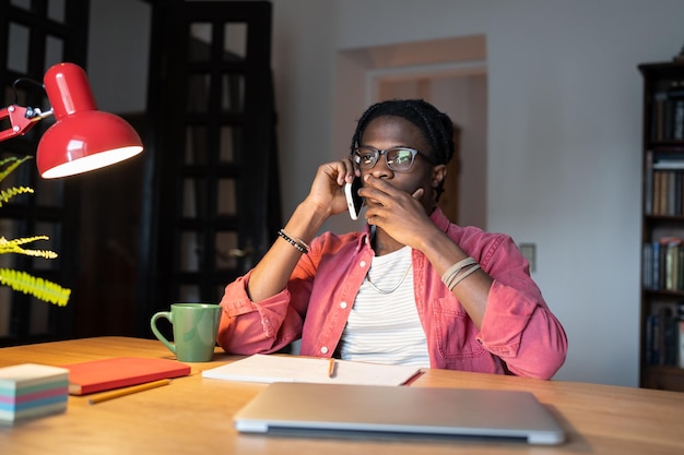 Gestresster, verwirrter afroamerikanischer Freiberufler, der telefoniert, sitzt mit Laptop und Tasse am Tisch