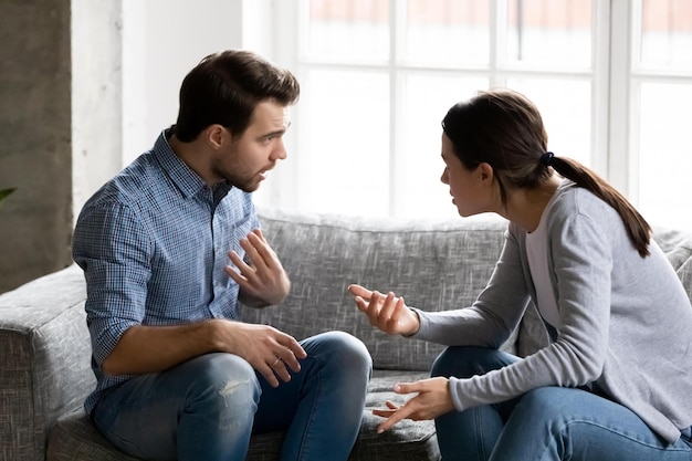 Gestresstes junges Ehepaar streitet sich emotional und beschuldigt sich gegenseitig, auf der Couch zu sitzen. Depressiver Ehemann streitet mit Ehefrau, die ernsthafte Kommunikationsprobleme hat