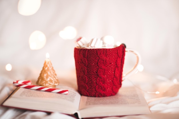 Gestrickte Tasse Kaffee mit Weihnachtssüßigkeit und Kerze auf offenem Buch über Lichtern im Zimmer. Weihnachtsferien. Wintersaison.