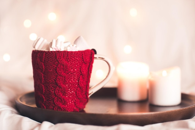 Gestrickte Tasse mit Kerze auf Holztablett hautnah über Weihnachtsbeleuchtung. Wintersaison.