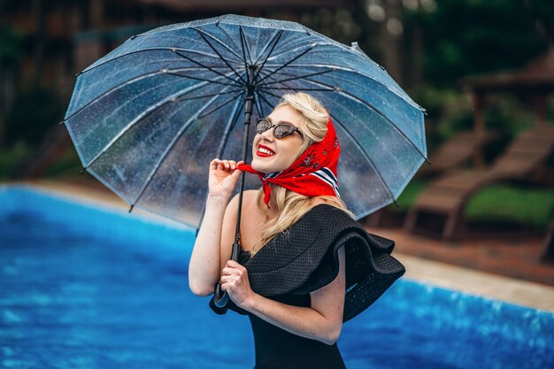 Gestylte Blondine mit Sonnenbrille, die einen Regenschirm im Schwimmbad hält