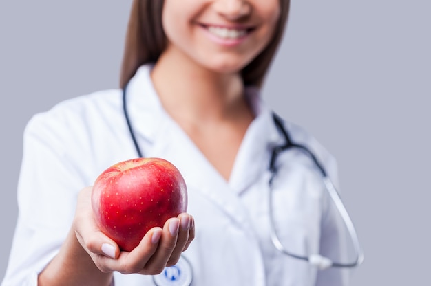 Gesund essen! Nahaufnahme der Ärztin in weißer Uniform, die roten Apfel hält und lächelt, während sie vor grauem Hintergrund steht