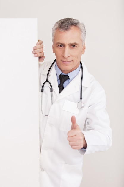 Gesund sein! Älterer Arzt mit grauem Haar in Uniform, der aus dem Kopierraum schaut und gestikuliert, während er isoliert auf weiß