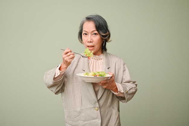 Gesunde 60er Jahre alte Frau, die gesunden grünen Salat isst, der isoliert auf grünem Hintergrund steht